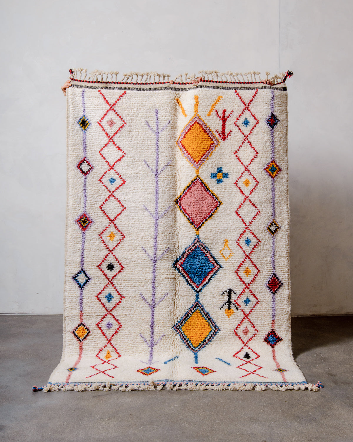 Berberlin Beniourain rug in colourful wool and cream, being held up in front of a white wall.